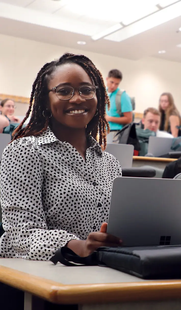 student in classroom smiling with laptop, other students visible in background