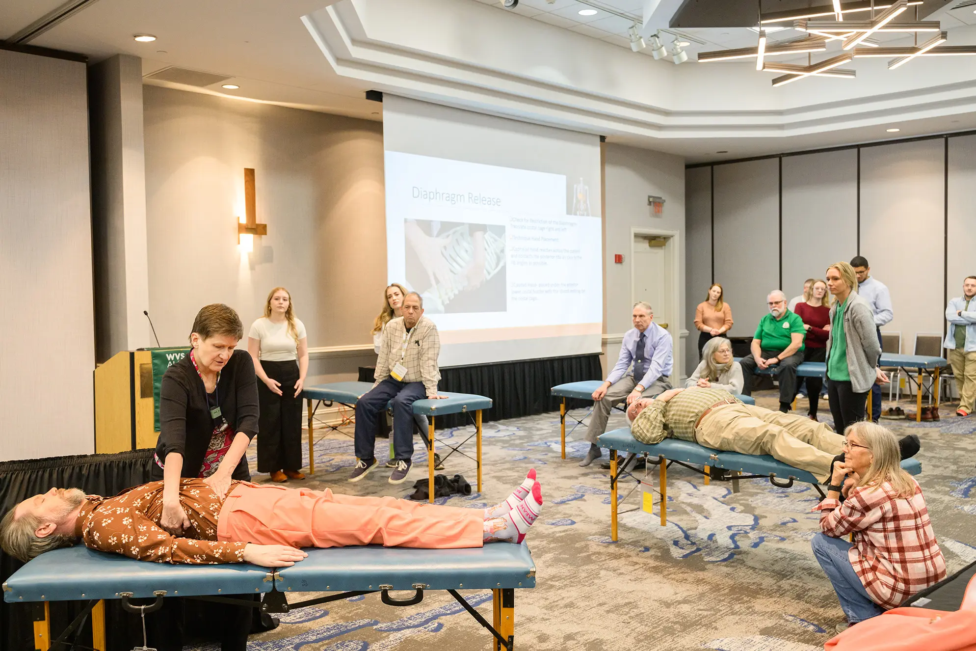 Two people lay on medical exam tables in a classroom while one is being observed. Onlookers watch the procedure.
