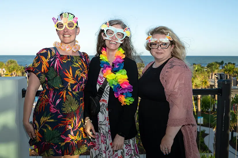 Three women smile for camera wearing silly glasses outside with ocean in background.