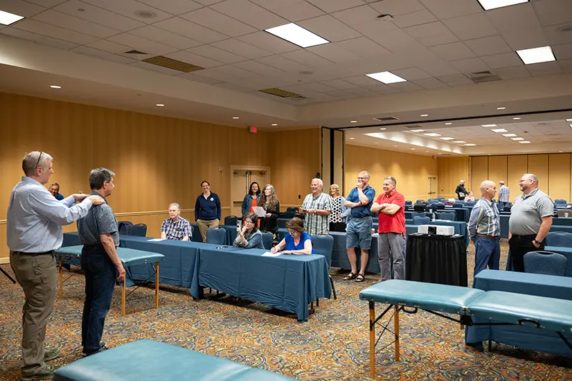 One man has both hands on another mands shoulders from behind. The two are in the front of an instructional class where others are standing watching. Seems to be demonstrating a procedure.