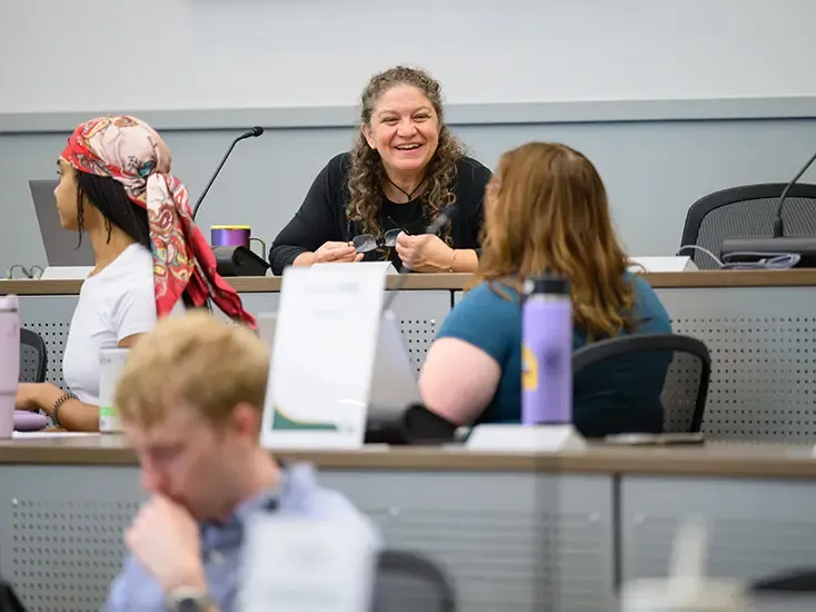 Students sitting in classroom turned speaking with professor seated one row behind them in tiered lecture hall.