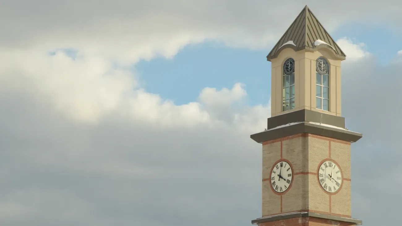 Alumni clock tower in foreground with clouded sky showing blue peaking through the center directly behind the tower.