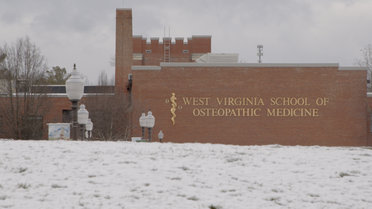 West Virginia School of Osteopathic Medicine displays on the side of brick building visible beyond a field of snow covered grass