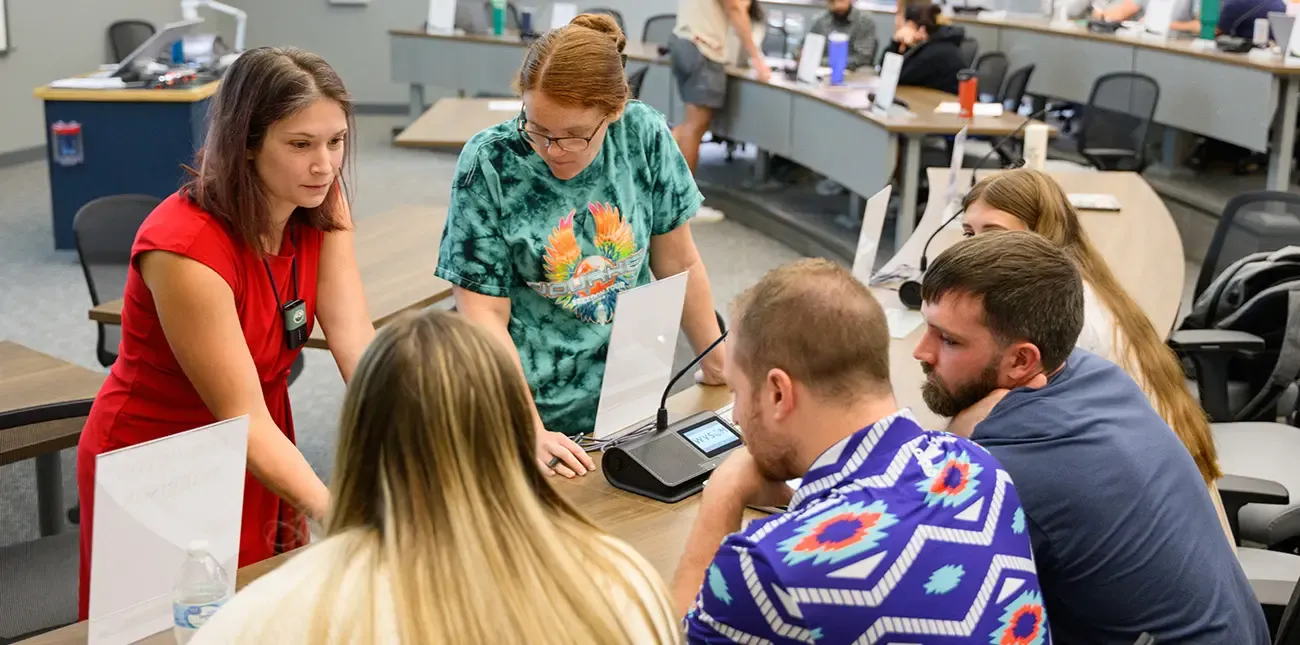 Instructor in classroom with students. Instructor close up to desk showing something to students that is blocked from view. Other students seen in background.