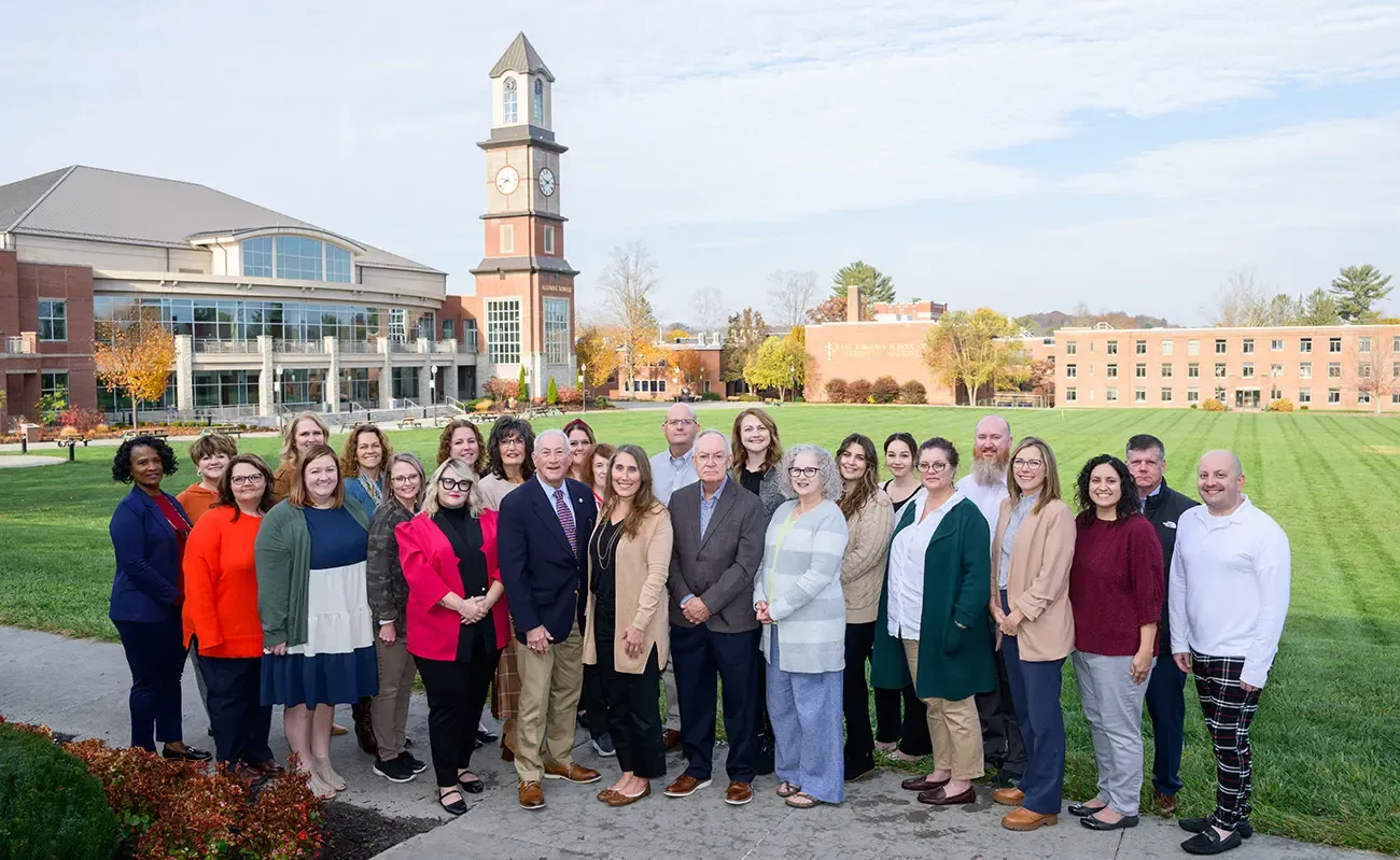 Group of people stand and smile for the camera outdoors during sunny day.  Student Center with Alumni Tower visible in background.