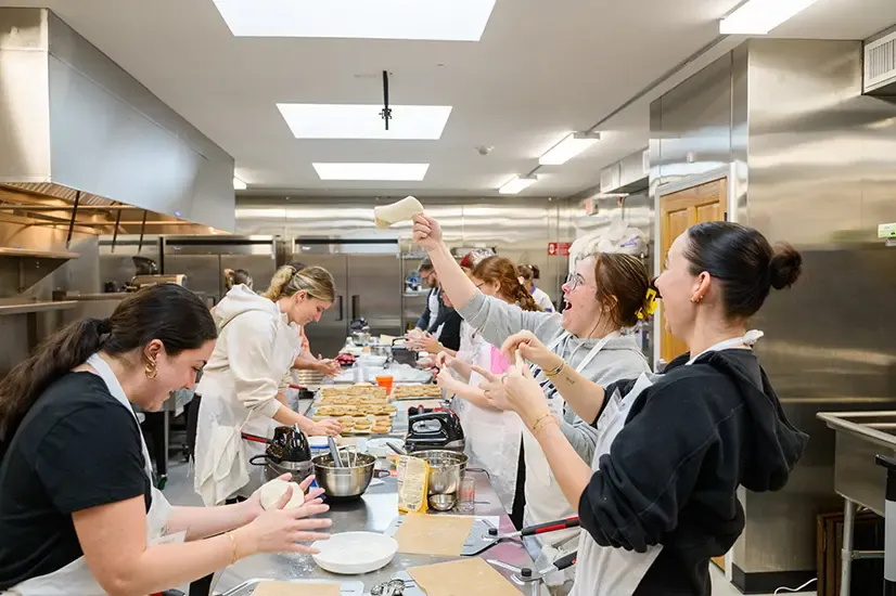 students in kitchen on left and right of prep table, one can be seen holding dough above head on outstretched finger, mood seems to be joyous.