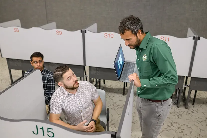 Students sit in individual numbered cubicles looking up and man holding a laptop. Main with laptop appears to be assessing something out of frame on the desk.