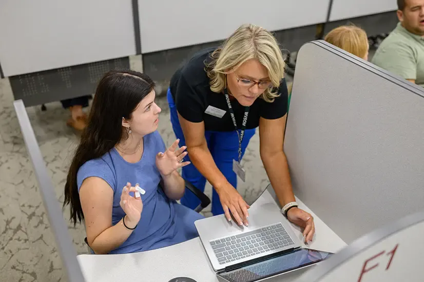 Woman wearing name tag helps student seated in cubicle with computer. 